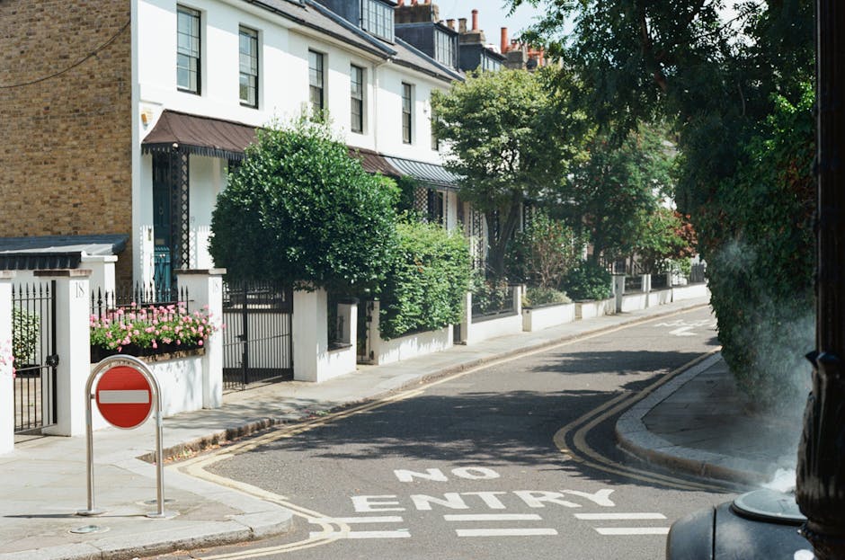 A narrow residential street with white townhouse-style buildings featuring black window frames and awnings, lush green trees and shrubbery lining the sidewalk, and a vibrant flower arrangement of pink