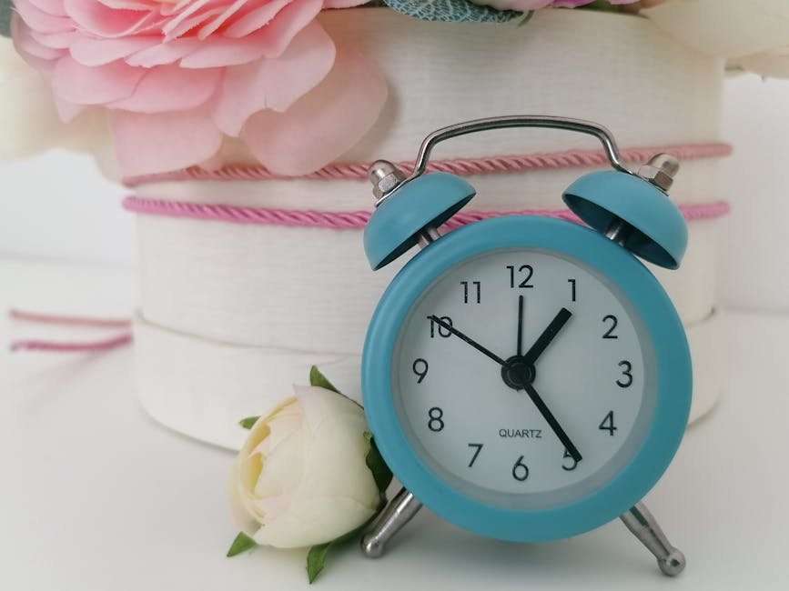 A light blue vintage-style alarm clock with twin bells and metal legs, showing the time as 12:05, is placed on a white surface near a white rose with green leaves. In the background, partially visible