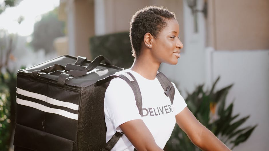 A young woman with short, curly hair wearing a white t-shirt with the word 'DELIVER' printed on it and a black backpack, standing outside during daylight. She is facing to the right with a gentle smil