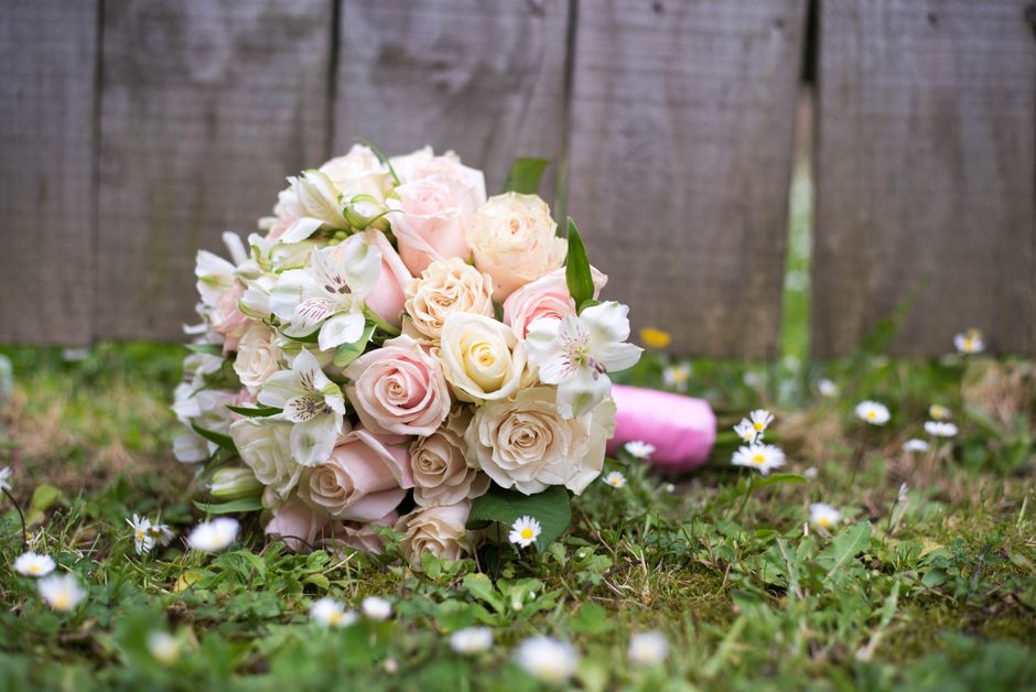 A bouquet arrangement of fresh, pastel-colored roses and white lilies, carefully placed on a grassy surface outdoors, with a wooden fence in the background. The roses feature soft pink and cream hues 