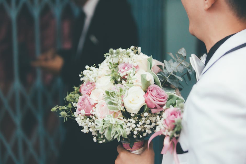 A man dressed in a white suit with a black trim is holding a floral bouquet in his hands, which features a mix of white and pink roses, small white baby's breath, and variegated green and white foliag