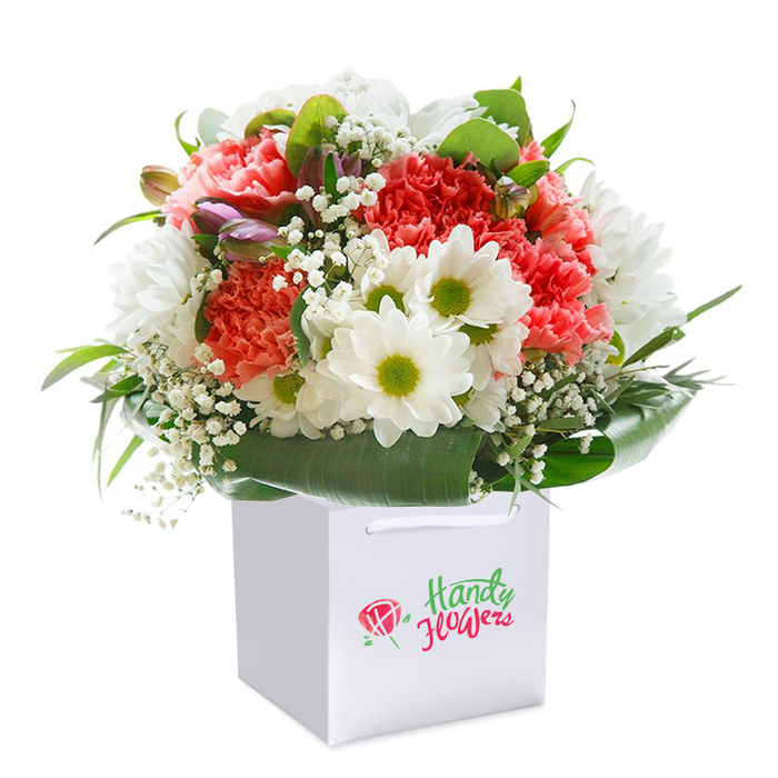 Rising elegantly from a crisp white square gift bag, this floral arrangement feels like a burst of refreshing colour in Marks Gate-perhaps echoing the vibrancy seen at the local markets or in the gardens dotting the neighbourhood. Plush coral and peach-hued carnations dominate the bouquet, their distinctive ruffled petals inviting closer inspection, while white daisy blooms with lively, lime-hued centers weave throughout, bringing a balance of brightness and cheer. Threads of white baby's breath interlace the ensemble, their delicate, airy structure softening the look and imparting a subtle sense of innocence. Look closely and you'll find a few closed buds tinged with purple nestled on the left edge, hinting at florals still to greet the day. Rounding the bouquet, rich green foliage with a waxy gleam makes a statement, both framing and protecting the more delicate blossoms. The rounded, abundant design is enhanced by careful placement and choice of blooms, each flower angled thoughtfully to catch the gentle, ambient light. Isolated against a pure white backdrop, the bouquet's composition, textures, and freshness are unmistakable, testifying to the skill of local Marks Gate florists. Perfect as a birthday gift, anniversary surprise, or a simple token of appreciation, this arrangement shines with the promise of joy and heartfelt connection.