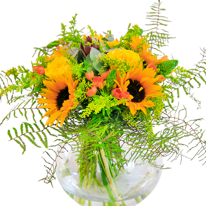 Upon first glance, this stunning bouquet captures the radiant essence of a late summer morning in Marks Gate, gathered by skilled hands at Marks Gate Florist. The focal point is an array of commanding sunflowers, their brilliant golden petals arching outwards to reveal chocolate-brown centers brimming with seed-each bloom basking as if in the glow of a Chadwell Heath sunrise. Nestled amongst them, garden roses unfurl in soft yellow spirals, their fullness conjuring images of cottage gardens along Rose Lane, providing a gentle, classic balance to the arrangement's vibrant heart. Peppering the bouquet, glossy red Hypericum berries glimmer with almost gem-like intensity, scattered across a medley of lush greenery. Feathery fern fronds and loose clusters of pale green filler weave through the bouquet, creating a lightness that recalls the easy breeze across the local parks. A few silvery eucalyptus leaves poke through, introducing subtle color shifts and aromatic freshness. The rounded glass vase, clean and simple, showcases the tangle of green stems reaching deep into cool, clear water-reminding onlookers of the vital beauty in every detail. This arrangement feels alive and inviting, its joyful color story and textured layers designed to brighten Marks Gate homes on birthdays, anniversaries, or spontaneous celebrations, embodying the community's warmth and cheer.