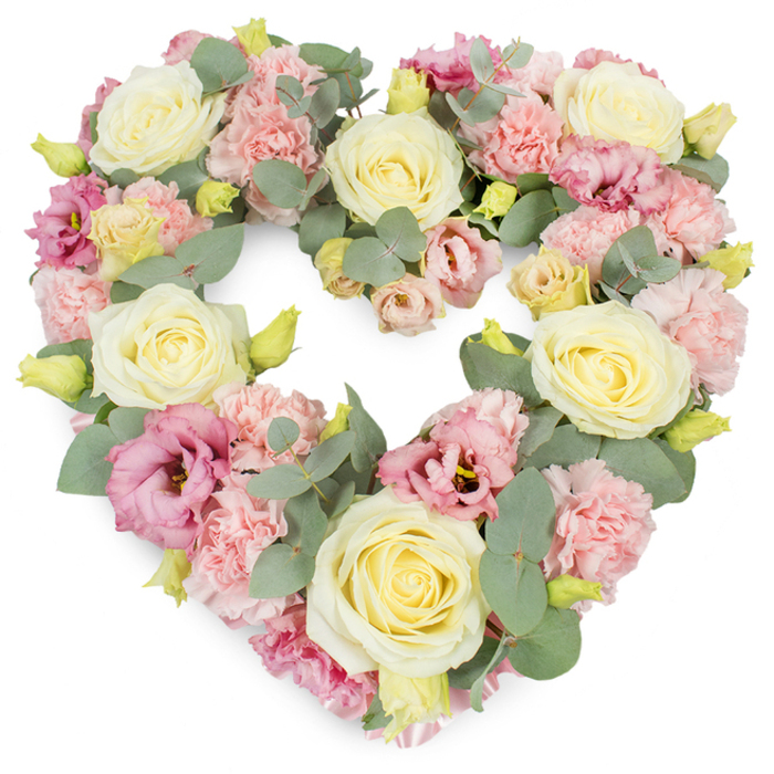 Against a perfectly white background, a luxurious heart-shaped wreath unfolds in soft, soothing tones, the kind of arrangement a local Marks Gate florist might prepare for a cherished farewell or memorial gathering. The heart shape is unmistakable and complete, traced by a dense mix of flowers and foliage so that the eye easily follows every curve. Cream and pale yellow roses appear at intervals across the wreath, their velvety petals curling from tight centres into open, rounded blooms that catch the light in gentle highlights and shadows. These roses lend a sense of poise and stability to the overall design. Clustered around them, pastel pink carnations provide volume and a rich, ruffled texture; each blossom is composed of many fine, crimped petals that seem to cushion and support the larger roses. Deeper pink to mauve lisianthus are scattered throughout, their more open, cup-like blooms and occasional darker centres adding depth and a soft transition between shades. Woven through the flowers, sprigs of silvery-green eucalyptus bring a calm, muted green tone, their round leaves creating a natural rhythm and soft structure. Here and there, tiny yellow-green buds are visible, suggesting the promise of new blossom and adding a fresh note to the tribute. The palette of cream, pink, and green is understated yet emotionally resonant, evoking peace, sympathy, and remembrance-ideal for funerals, memorials, or anniversaries in and around Marks Gate, Collier Row, or Chadwell Heath. One can almost imagine the gentle mix of floral and eucalyptus fragrance accompanying a quiet service, the heart wreath standing as a tender symbol of affection, gratitude, and the unbroken bond with someone dearly missed.